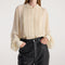 A woman wearing a cream-colored long-sleeve blouse with pleated front detailing and a scalloped collar, paired with black pants, against a plain light background.
