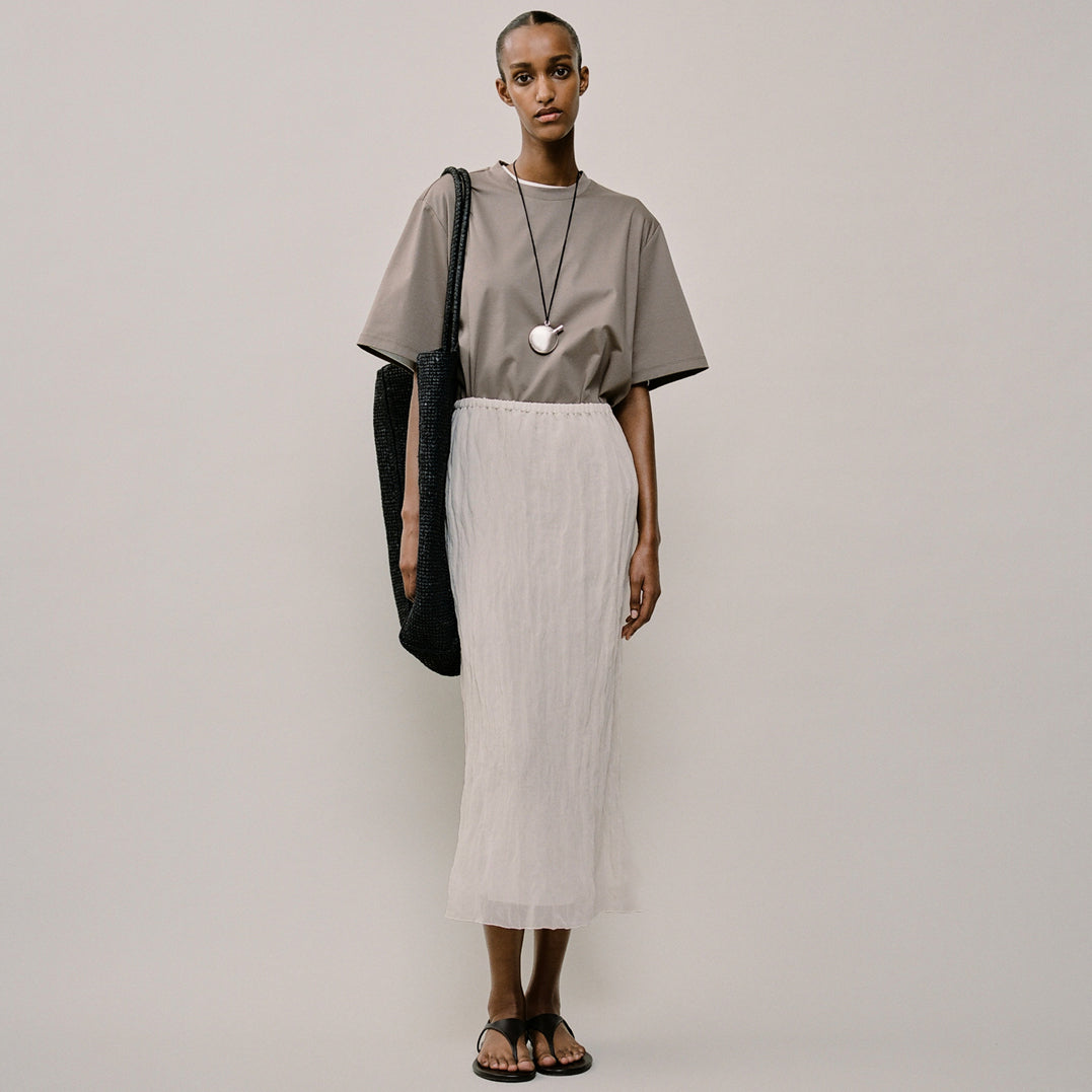 Woman wearing a gray top and white skirt with a black bag on a plain background