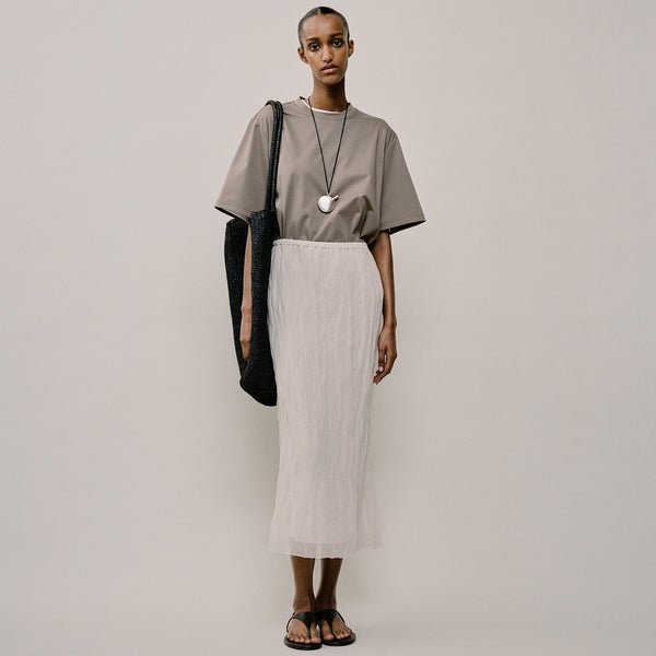 Woman wearing a gray top and white skirt with a black bag on a plain background